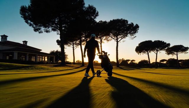 Silhouetted golfer walking at sunset pulling golf trolley across fairway near clubhouse