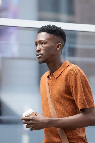 African man walking urban street wearing rust-orange polo holding coffee cup and crossbody bag