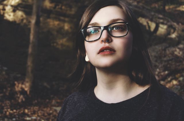 Young woman with glasses and piercings standing outdoors surrounded by trees and leafy ground cover. She looks confident and serene. Perfect for use in lifestyle, fashion, or outdoor-themed projects.