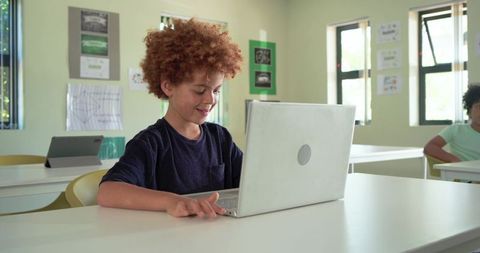 African schoolboy using laptop in modern classroom smiling learning with tablet nearby