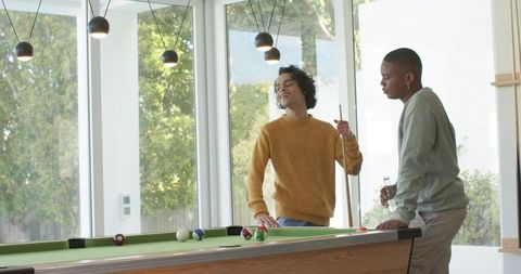Young men laughing and playing pool in sunlit modern home with cues and pendant lights