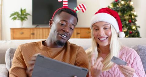 Diverse Couple Online Shopping for Christmas on Sofa with Festive Headwear