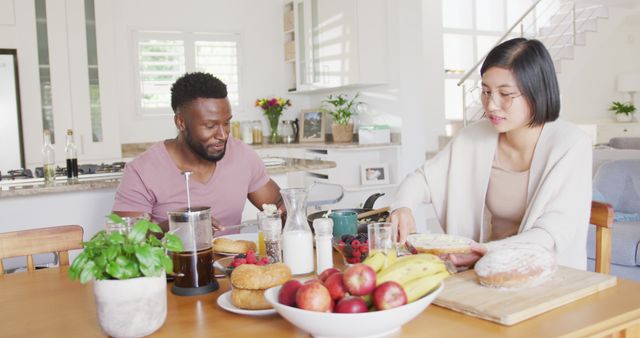 Depicts a multiethnic couple enjoying breakfast in a bright and cozy home kitchen. Ideal for use in lifestyle blogs, health and wellness articles, and promotional material for kitchen accessories. Emphasizes themes of togetherness, diversity, and healthy living.