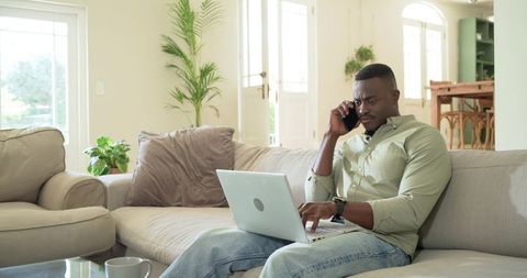 Mid-adult african american man working on laptop and talking on phone on sunlit sofa