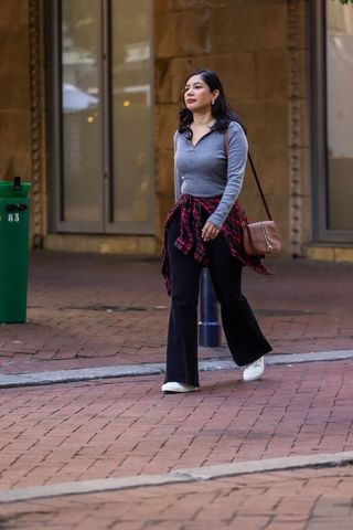 Woman walking on brick sidewalk wearing plaid shirt tied at waist with brown crossbody bag