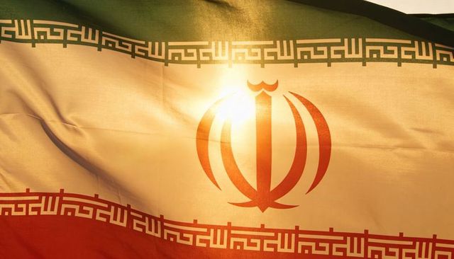 Waving iranian flag with central emblem and kufic border, backlit sunflare on fabric