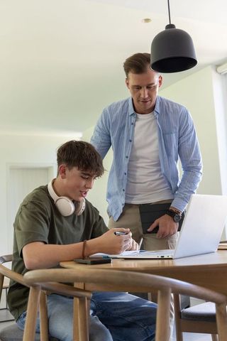 Father and teenage son studying at home at wooden table with laptop and notebook