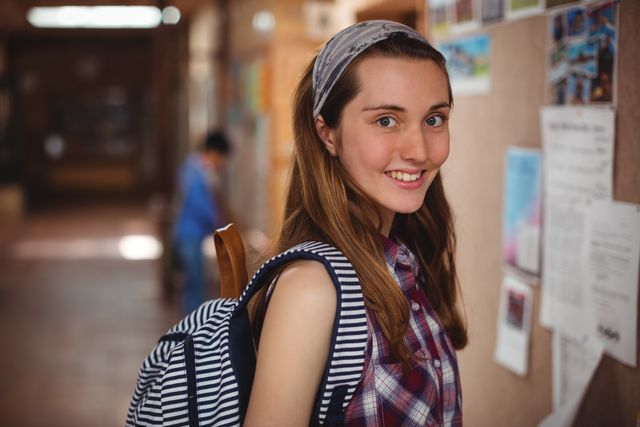 This image shows a cheerful schoolgirl standing near a notice board in a school corridor. She is wearing a striped backpack and a plaid shirt, giving a casual and approachable vibe. This image can be used for educational websites, school brochures, student life articles, and promotional materials for academic institutions.