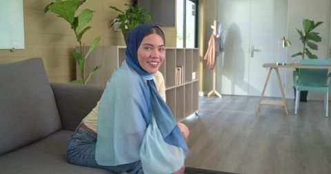 Indian woman wearing blue hijab smiling while sitting on sofa in modern living room
