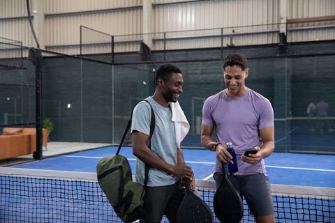 Smiling diverse male friends at padel court net holding paddles, bottle and phone