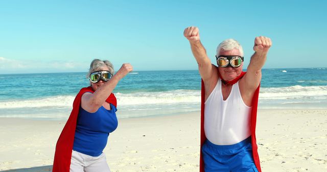 Image of a senior couple dressed as superheroes with red capes and goggles, standing on a beach near the ocean. Use this for themes of active aging, fun in retirement, enjoying life, and elder adventures. Perfect for advertisements or media related to travel, health, senior living, and lifestyle.
