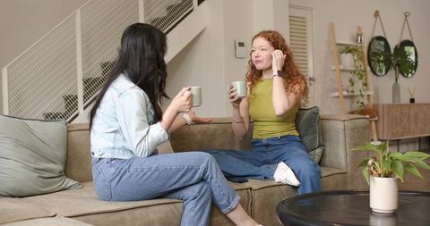 Diverse friends sitting on couch drinking coffee and chatting in modern cozy living room