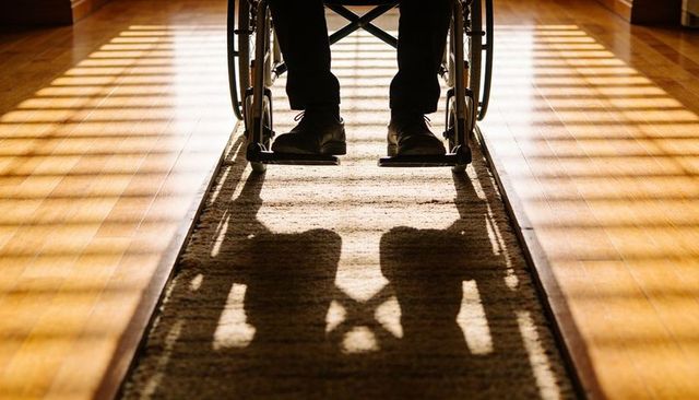 Wheelchair user casting dramatic shadow on sunlit hardwood corridor rug