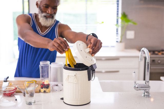 Senior african american man putting banana peel in small dustbin on kitchen counter at home. unaltered, people, waste management and responsibility concept.