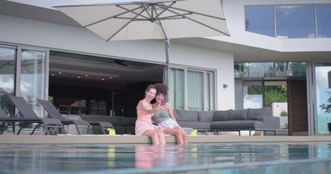 Two diverse women smiling and taking selfie while sitting on luxury poolside terrace