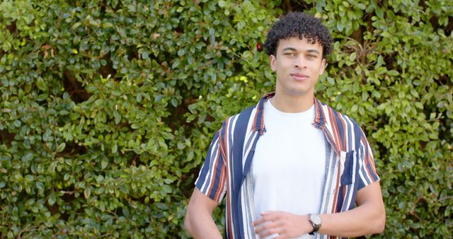 Young man standing against a background of green foliage, dressed casually, and smiling. Great for promoting outdoor adventures, fashion brands, relaxation products, and lifestyle blogs.