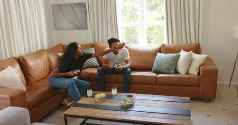African american couple playing video games on leather sectional sofa in living room