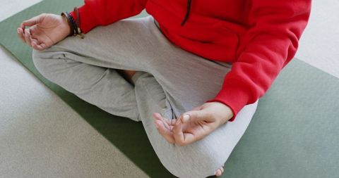 Male practicing mindful meditation on green yoga mat wearing red hoodie and gray pants