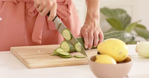 Woman Wearing Apron Slicing Cucumber on Cutting Board in Kitchen