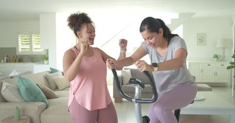 Diverse female friends cheering together pedaling stationary bike in bright living room