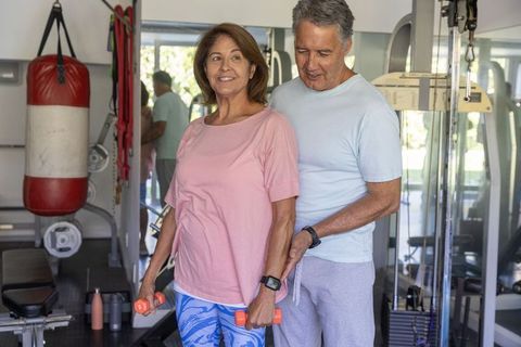 Senior couple exercising together in home gym while partner guiding dumbbell exercise