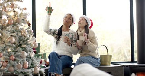 Diverse women wearing santa hats sipping hot cocoa and pointing at christmas tree