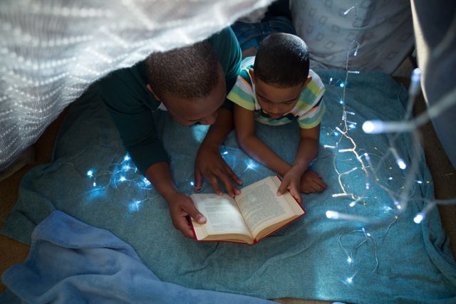 Father and son enjoying quality time reading a book together in a cozy fort adorned with fairy lights. Perfect for illustrating family bonding, bedtime routines, and the importance of reading and education. Ideal for use in parenting blogs, educational materials, and family-oriented advertisements.