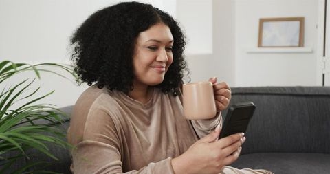African American woman in loungewear sipping coffee and scrolling smartphone on cozy sofa at home