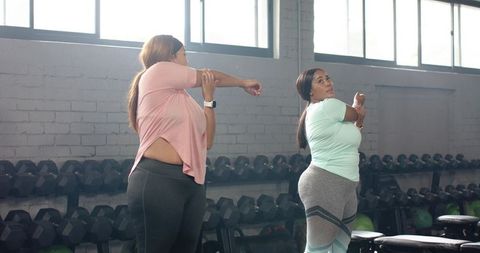 Plus-size women stretching together in gym by dumbbell rack, body positive fitness duo