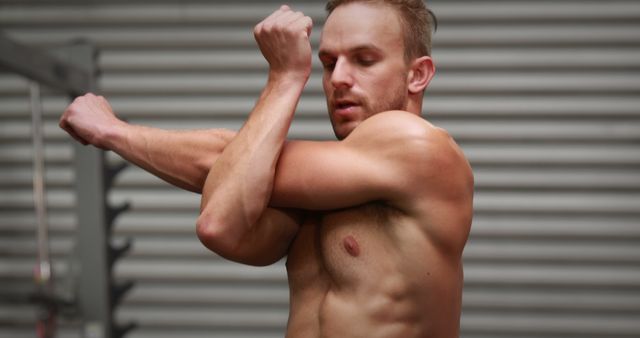 A shirtless young Caucasian man is stretching his arms before a workout, with copy space. His focused expression and dynamic pose suggest he is preparing for physical exercise or sports activity.