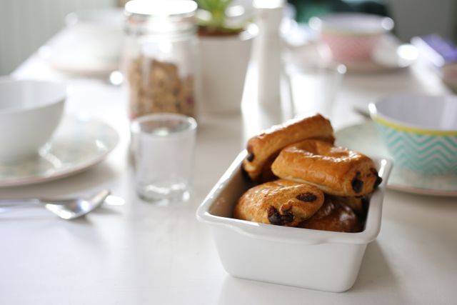 Freshly baked pastries sit in a white dish on a breakfast table. The table is set with bowls, glasses, and a jar of cereal, suggesting a wholesome morning meal. This can be used for images relating to breakfast, baking, home dining, and morning routines.