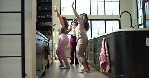 Diverse friends dancing in bright modern kitchen wearing loungewear and holding glasses