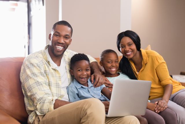 This image shows a joyful African American family sitting on a sofa at home, using a laptop together. The parents and children are smiling, indicating a moment of bonding and happiness. This image can be used for advertisements, articles, or websites related to family life, technology in the home, parenting, or leisure activities.