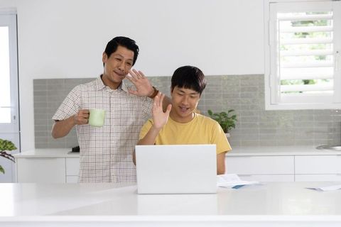 Asian father and son waving on video call at bright kitchen island, casual morning