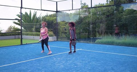 African american friends smiling playing padel on blue outdoor court with glass walls