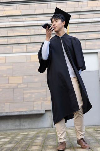 Asian male graduate speaking into smartphone on campus wearing gown and mortarboard