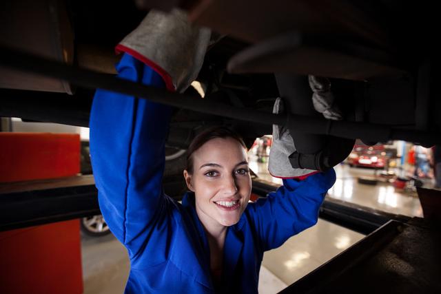 Smiling female mechanic in blue uniform servicing a car at a repair garage. Suitable for promoting automotive repair services, showcasing women in skilled trades, illustrating mechanic training programs, or featuring professional automotive workshops.