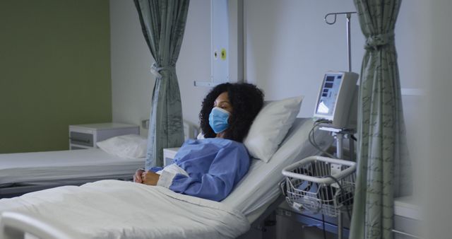 Young woman lying in hospital bed, wearing gown and mask, appears resting or recovering. Ideal for medical, healthcare, recovery, or pandemic-related themes, could be used by hospitals, clinics, or health publications to depict patient care and safety.