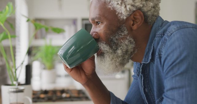 Elderly man with gray beard and casually dressed in denim drinking coffee in cozy bright kitchen. Ideal for articles and advertisements about senior lifestyle, morning routines, relaxation at home, and promoting beverages or kitchen products.