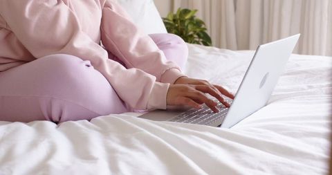 Woman typing on laptop in pink loungewear on white bed, cozy minimalist home workspace