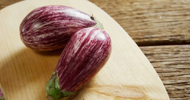Two heirloom eggplants with purple and white stripes lying on a light-colored wooden board atop a rustic wooden surface. Ideal for use in cooking blogs, health and wellness websites, farm-to-table restaurant promotions, and farmer's market advertisements illustrating organic produce and fresh vegetables.