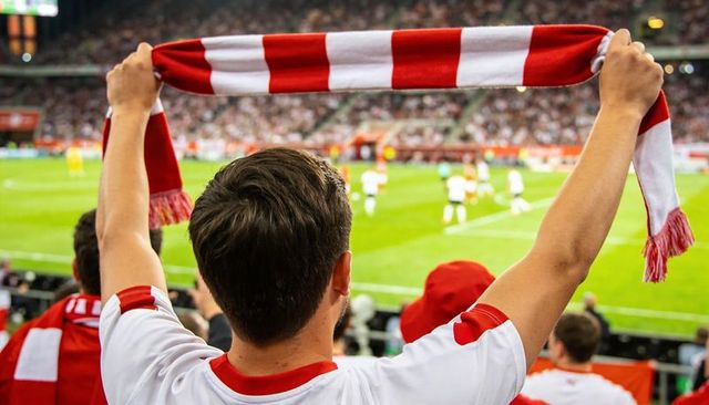 Male fan raising red and white scarf cheering and celebrating in packed soccer stadium