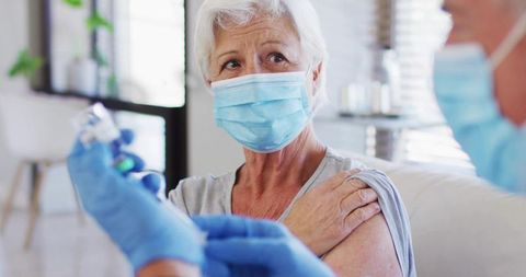 Senior woman receiving vaccine at home from masked caregiver wearing gloves during home visit