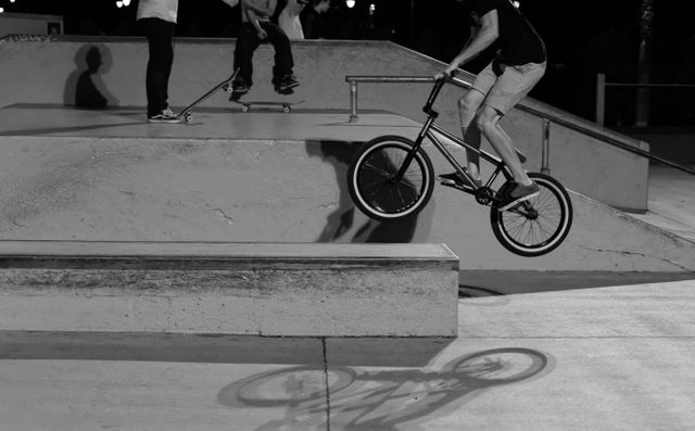 Teenage boy riding BMX bike and performing trick on ramp in skate park during nighttime. Shadow casts on concrete ground, showcasing dynamic movement and skill. Ideal for use in articles about extreme sports, urban youth culture, skateboarding and BMX activities, night photography.