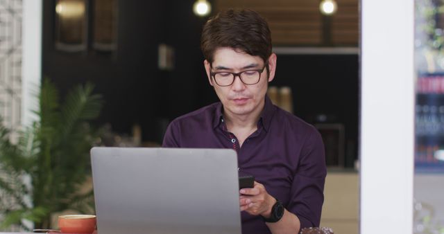 A man wearing glasses working on a laptop while checking his smartphone in an office. Ideal for use in business technology, remote work, productivity, and workplace efficiency themes.