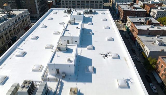 Drone hovering over white commercial rooftop with skylights and hvac units in downtown city