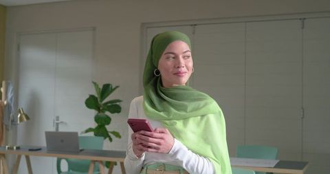 Asian woman wearing green hijab holding smartphone in home office with laptop, smiling