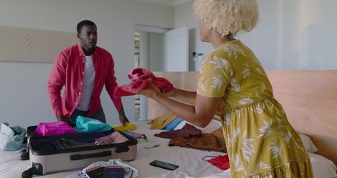 Diverse couple packing and sorting clothes on bed with open suitcase and passports