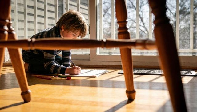 Sunlit boy drawing on hardwood floor near sliding glass door with colored pencils