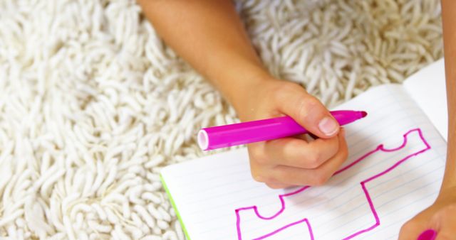 Child drawing various shapes with a pink marker in a notebook while lying down on a carpet. Ideal for educational, artistic, or child development themes. Useful for promoting creativity, homeschooling activities, or back-to-school scenarios.
